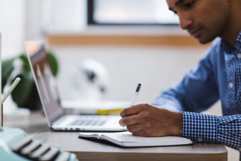 Homem faz anotações em um caderno ao lado do notebook aberto