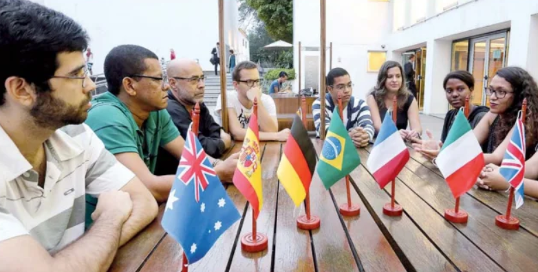 Membros do Clube Poliglota Brasil reunidos em torno de uma mesa com bandeiras de vários idiomas durante um encontro presencial